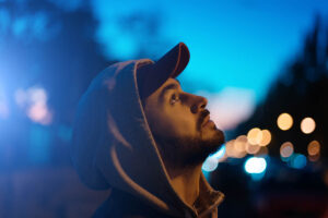 a man wearing a hoodie and a baseball hat in the dark looks up to the sky and asks himself how to overcome opioid addiction