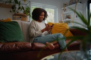 a woman sits on her couch looking at her phone searching for a mental health day treatment program that will suit her needs