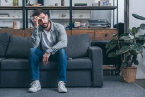 man sits on his couch looking distraught and considering that he might need to research susbtance abuse treatment programs