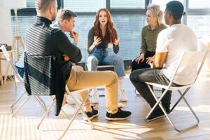 a group of five people sit in a circle participating in a group therapy session and discuss how addiction treatment programs can help them