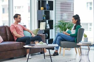 a man sits on a couch in front of a therapist sitting in a chair talking about the man's intensive outpatient program