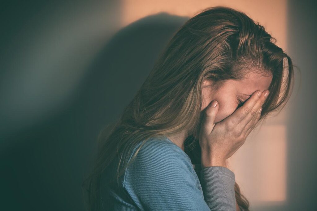 a woman stands with both palms to her face struggling with the signs of alcohol withdrawal