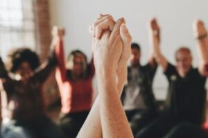 a big group of people sitting are raising and holding each others hands and benefiting from their 12-step treatment programs