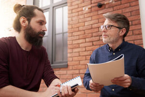 a man with beard talks to a man holding papers discussing with him the advantages of a men's rehab program