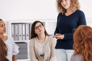 a group of women sit and listen to another woman standing talking about the benefits of a women's rehab program