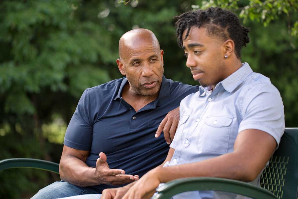 an older adult male sits on a bench with a younger adult male and talks about how addiction affects the family dynamic
