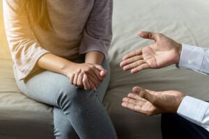a person sits on a couch with hands folded while her therapist their palms open explaining the benefits of one on one addiction counseling
