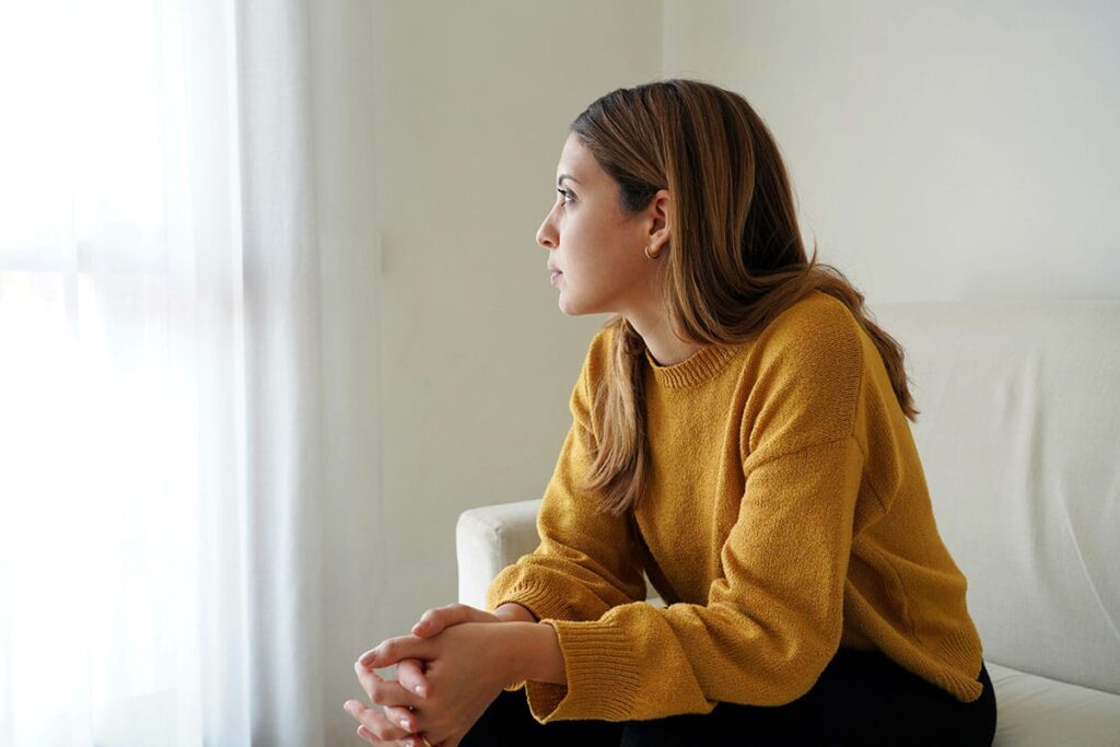 a young adult woman sits with hands clasped together looking at a window wondering how painkillers become addictive