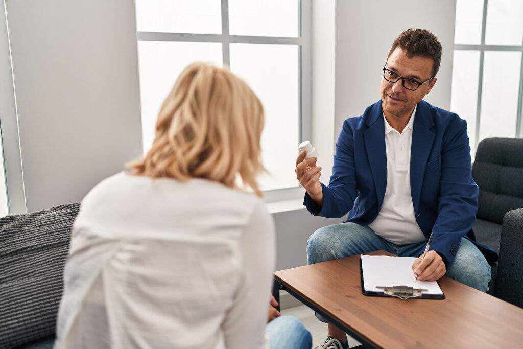 a man with glasses sits in front of a table with a clipboard on it and answers the woman sitting across from him question about what is mat