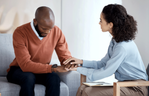 distraught man who is healing from trauma sits on a couch while his therapist holds his hand