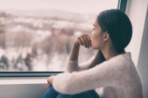 woman sits near a window and looks out the window while hold her hand not her mouth thinking about upcoming holiday stress