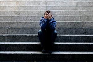 a person sits outside on steps and holds their hands to their head struggling with the signs of binge drinking