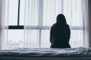 person sits at the edge of their bed looking out their window thinking about the long-term effects of ptsd