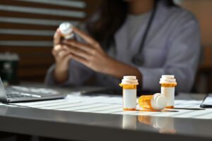 a medical professional holds a bottle of medicine while other medication bottles are the table and discusses the dangers of opioid dependence to patient