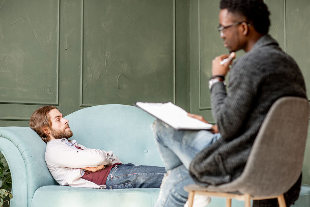 man sits on a couch and is talking to his therapist who has a clipboard and is explaining some dbt techniques