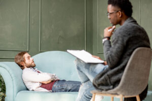 man sits on a couch and is talking to his therapist who has a clipboard and is explaining some dbt techniques